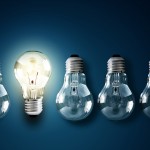 4 unlit, upside-down glass lightbulbs with one lit, right-side up bulb on a dark-blue, gradient background