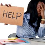 woman at computer covering her face with one hand and holding a cardboard sign that in the other that reads: help